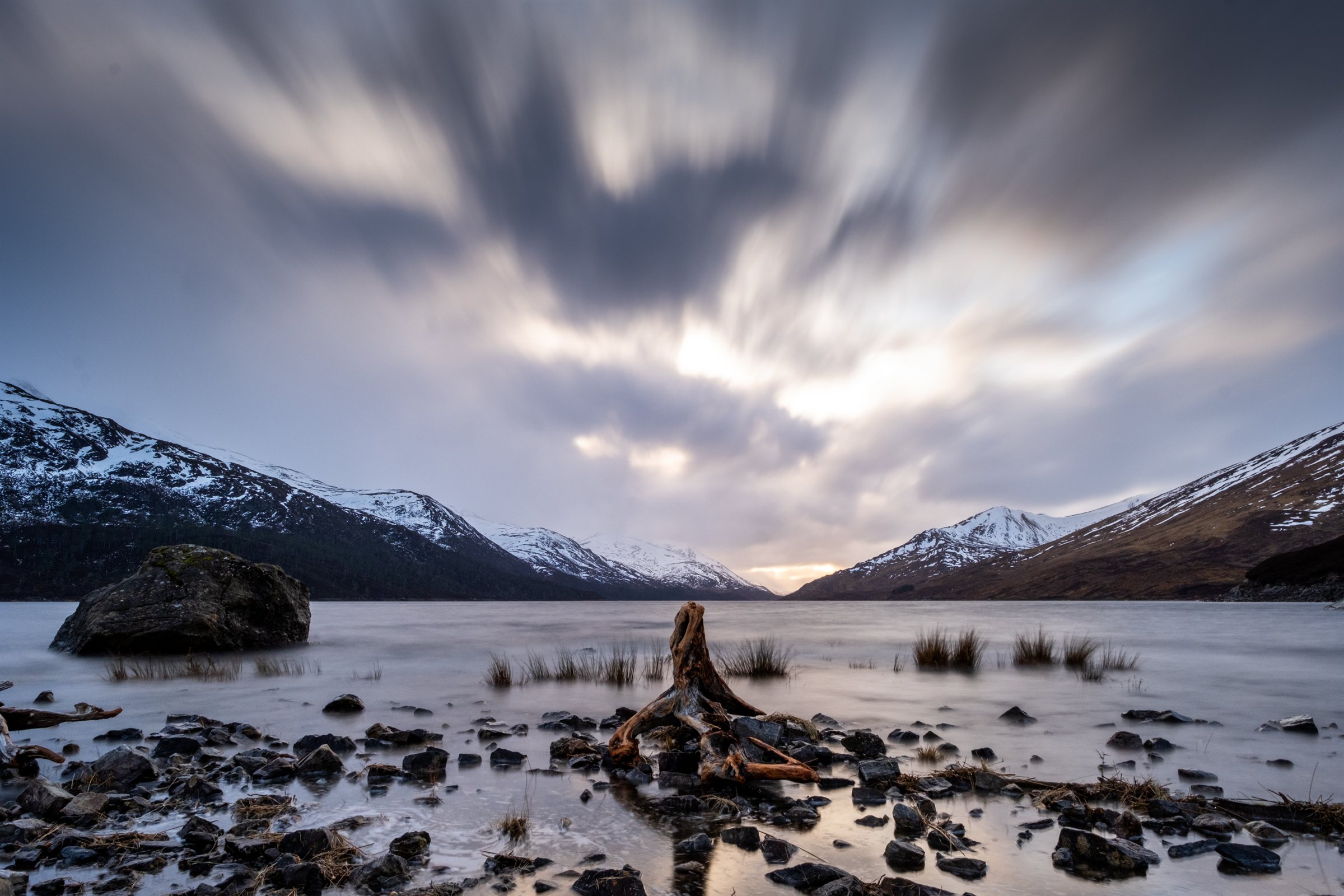 Rocks in a loch beneath snowy peaks and streaming cloud