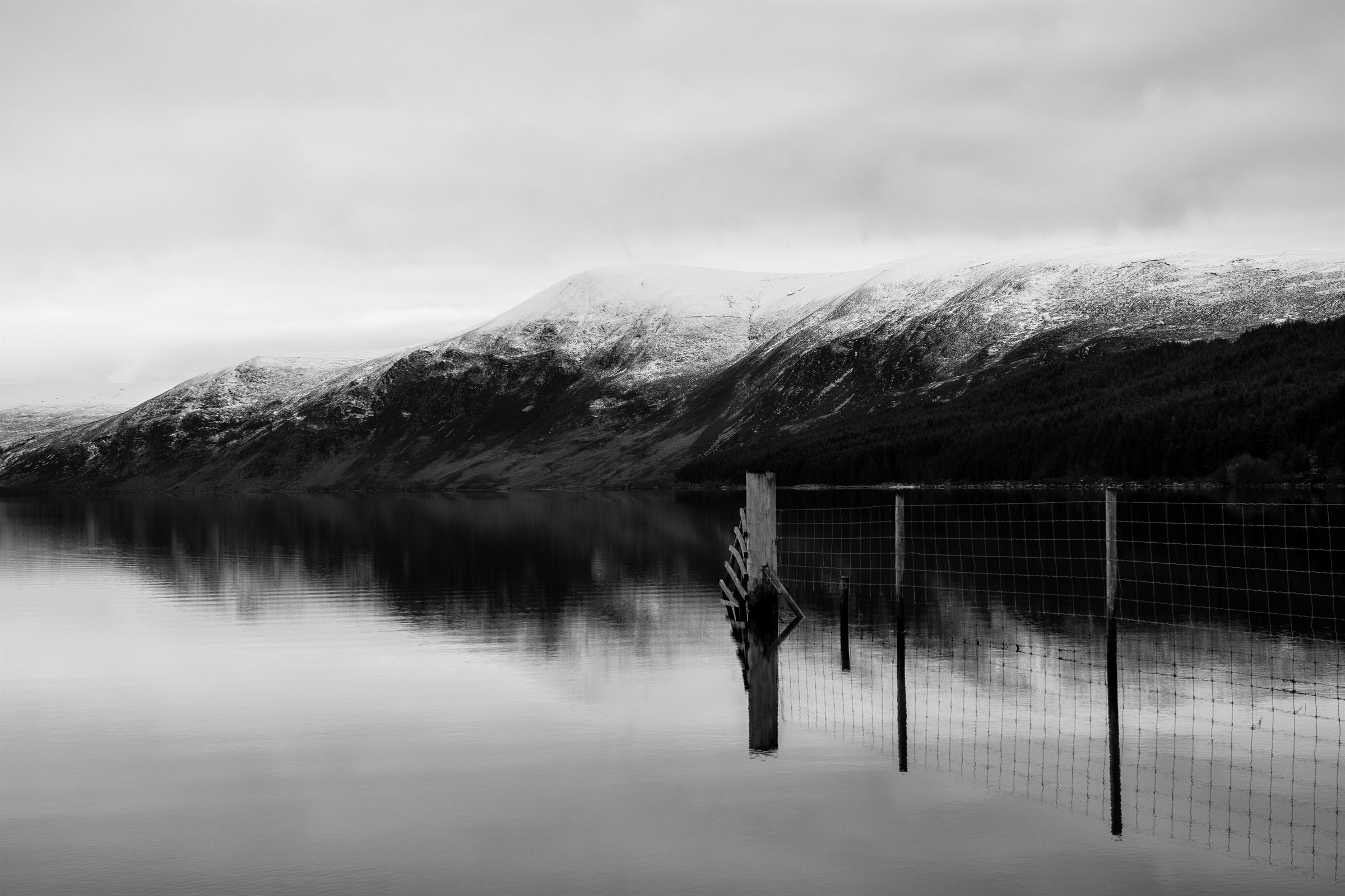 Snow-topped hills reflected in a still loch, an old fence running into the water