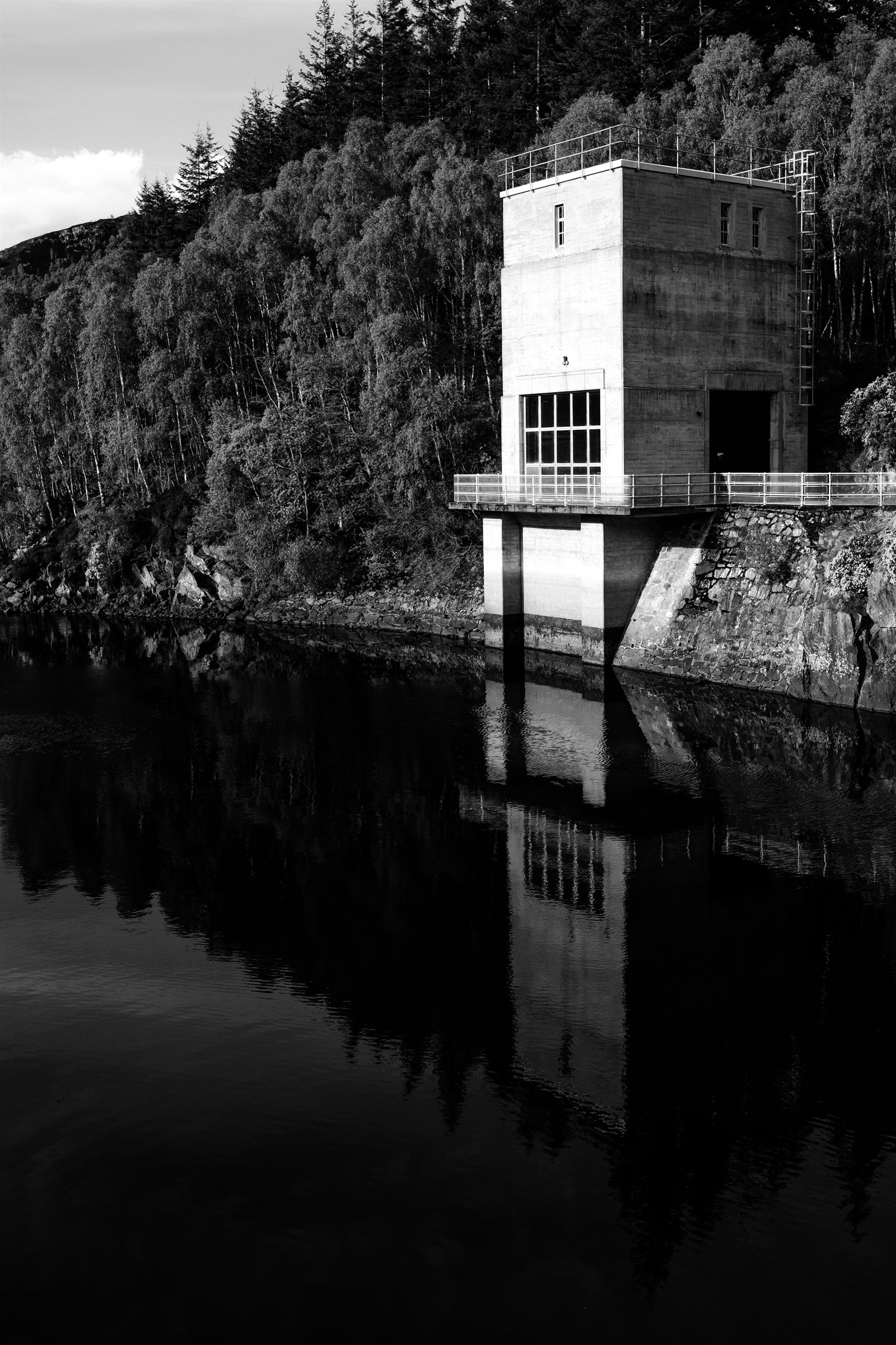 A concrete tower reflected in dark water beneath wooded hills