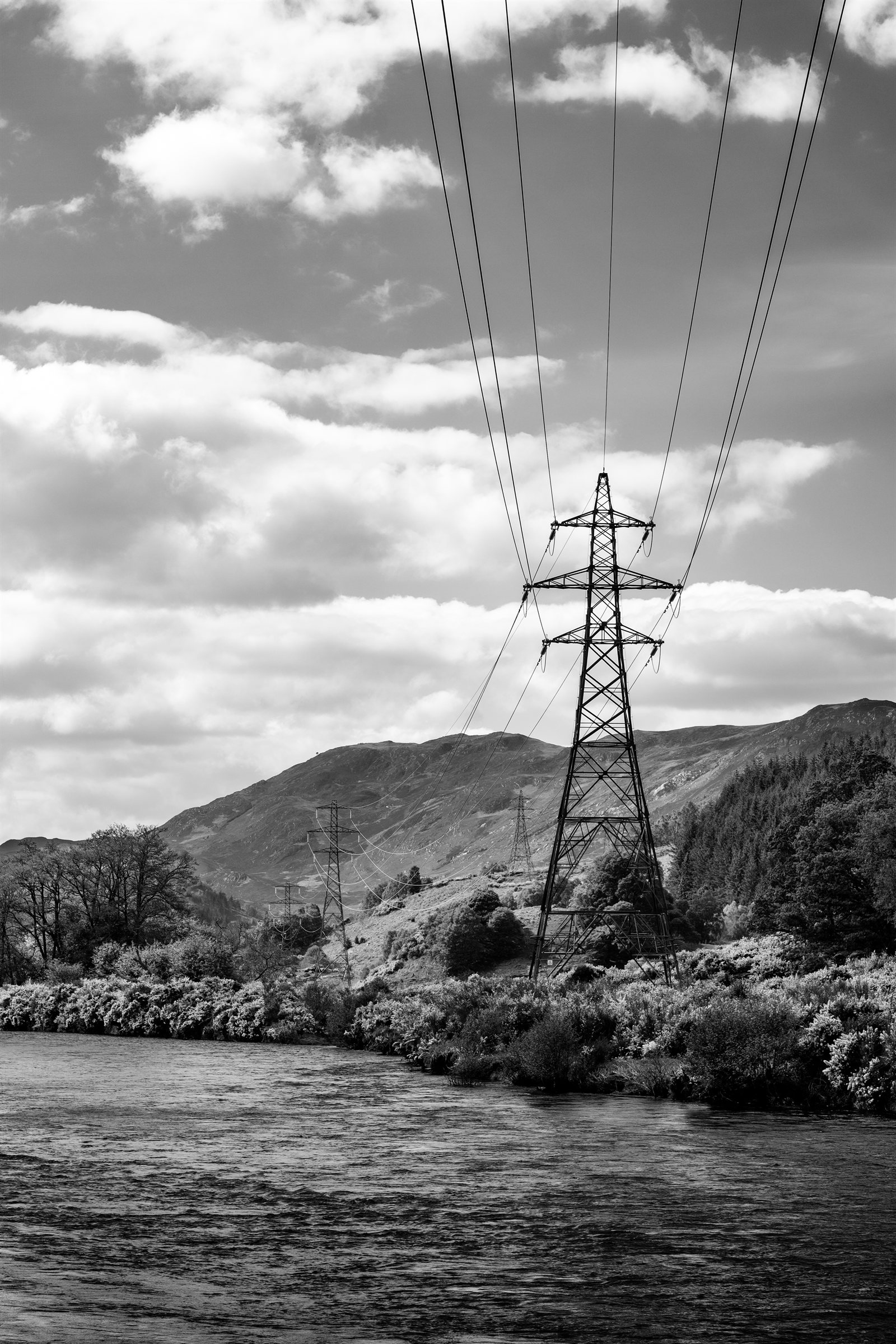A pylon above a river bank, wires running up into the clouds