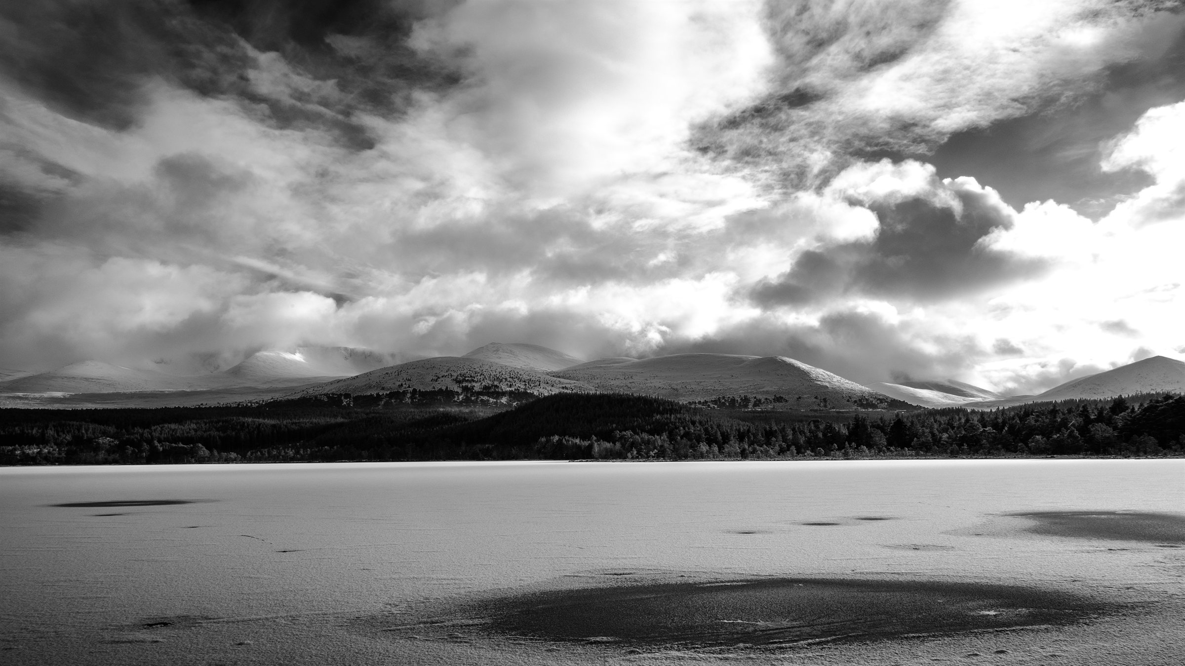 A frozen, snow-covered loch below forest and snowy mountains under a dramatic sky