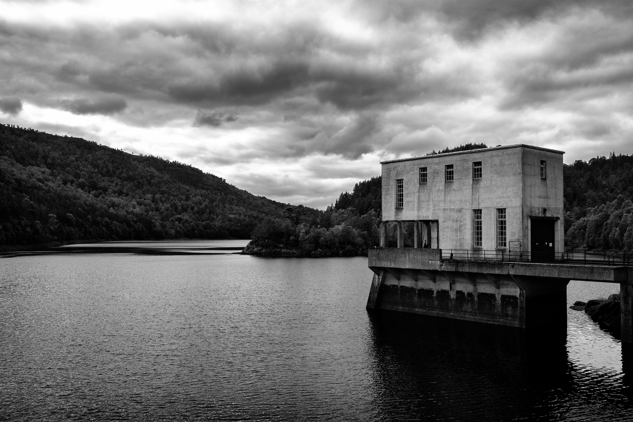 A concrete intake tower standing in a loch beneath wooded hills