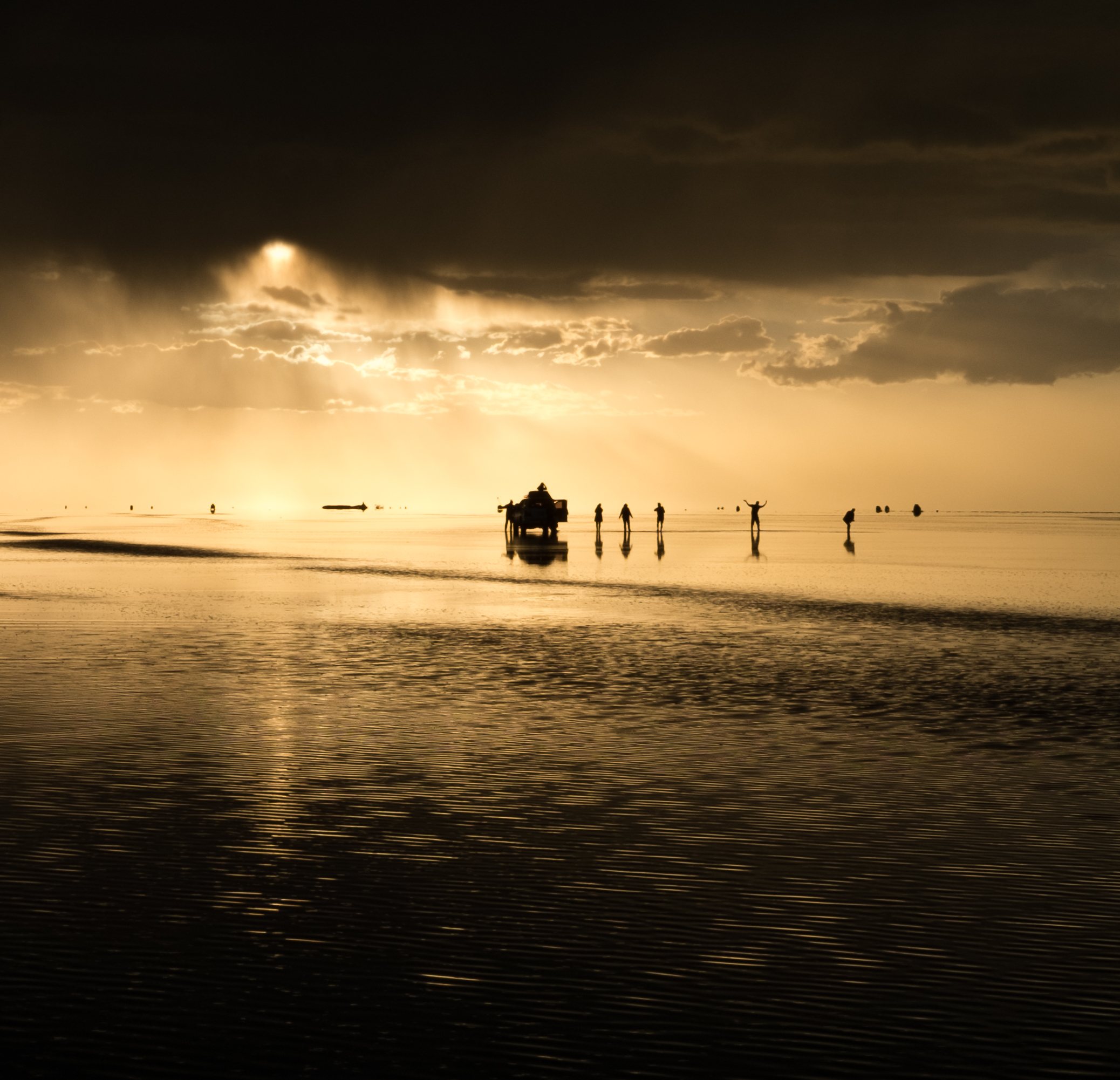 People and a jeep silhouetted on golden salt flats at sunset