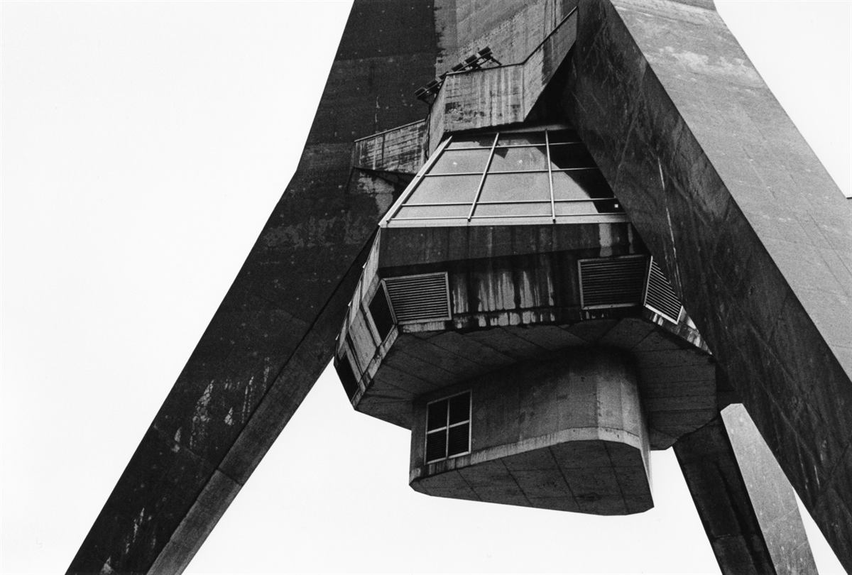 Looking up at a brutalist concrete tower and its suspended observation pod against a pale sky, in black and white