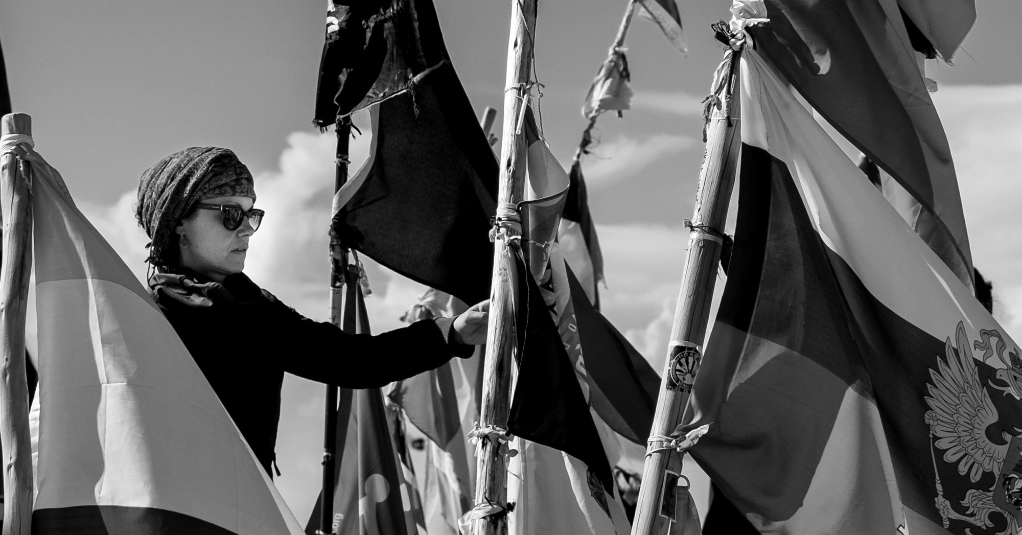 A person in sunglasses and a patterned headscarf reaching among a crowd of flags on wooden poles, in black and white