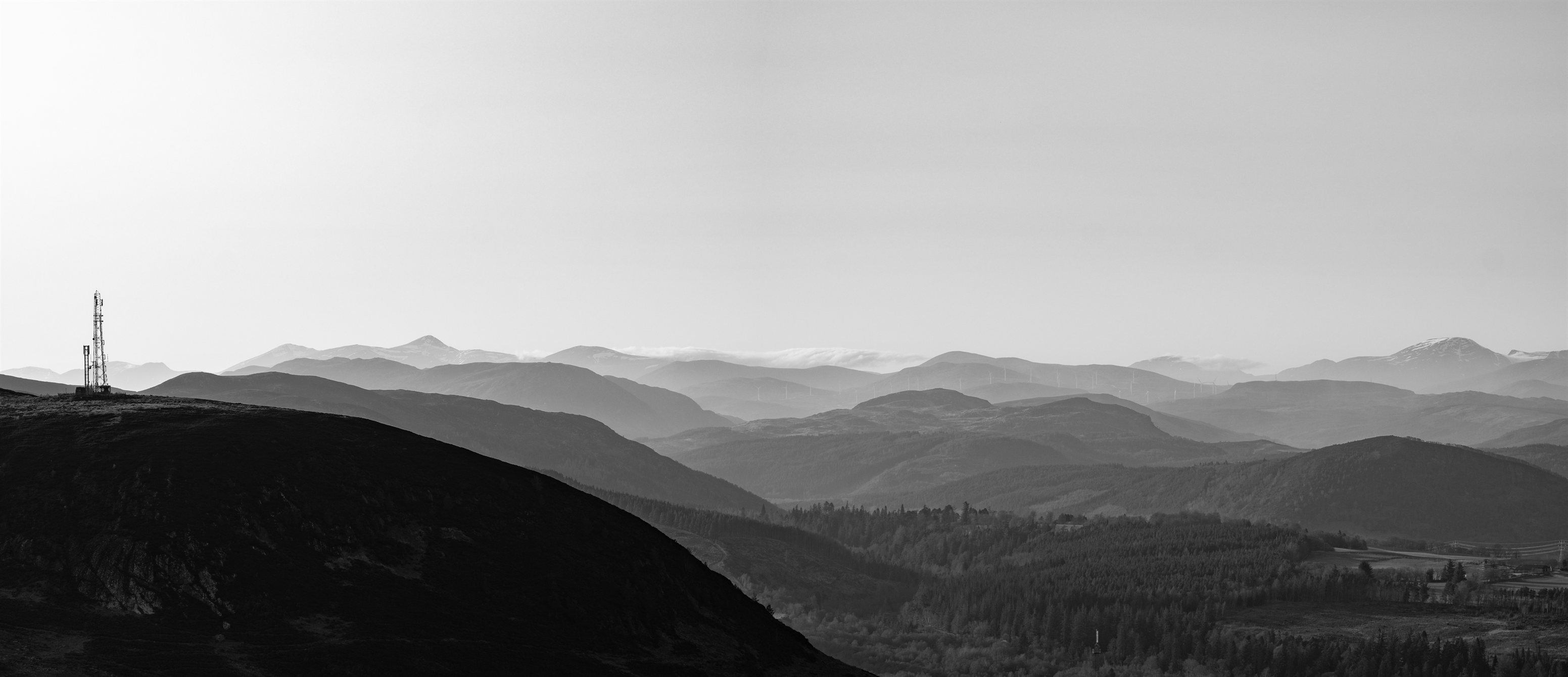 A wide panorama of layered hazy hills and distant snow-capped mountains, a radio mast on a dark hilltop at the left, in black and white