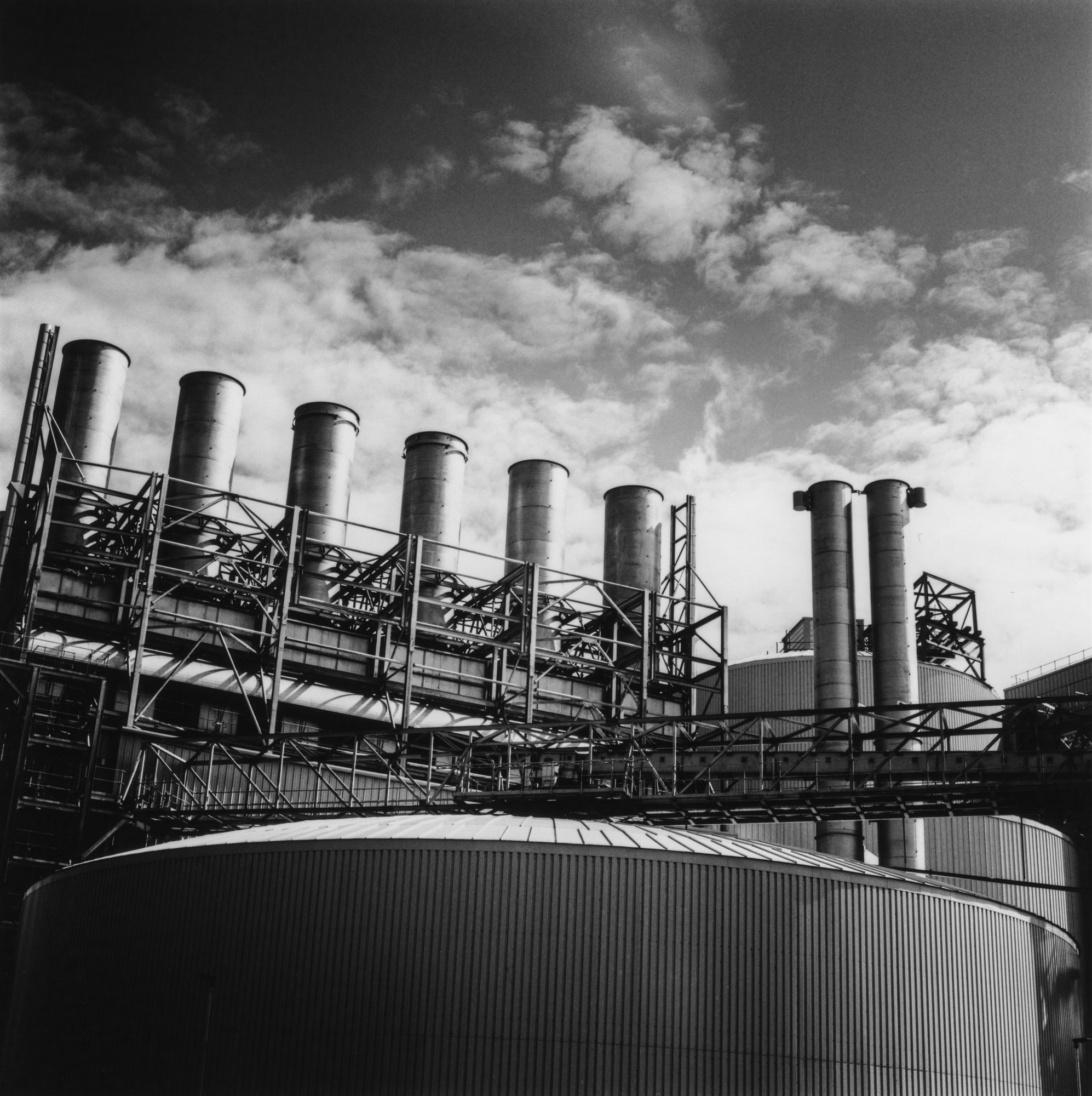 A row of steel chimneys and lattice walkways above a curved industrial tank, under a cloudy sky, in black and white film