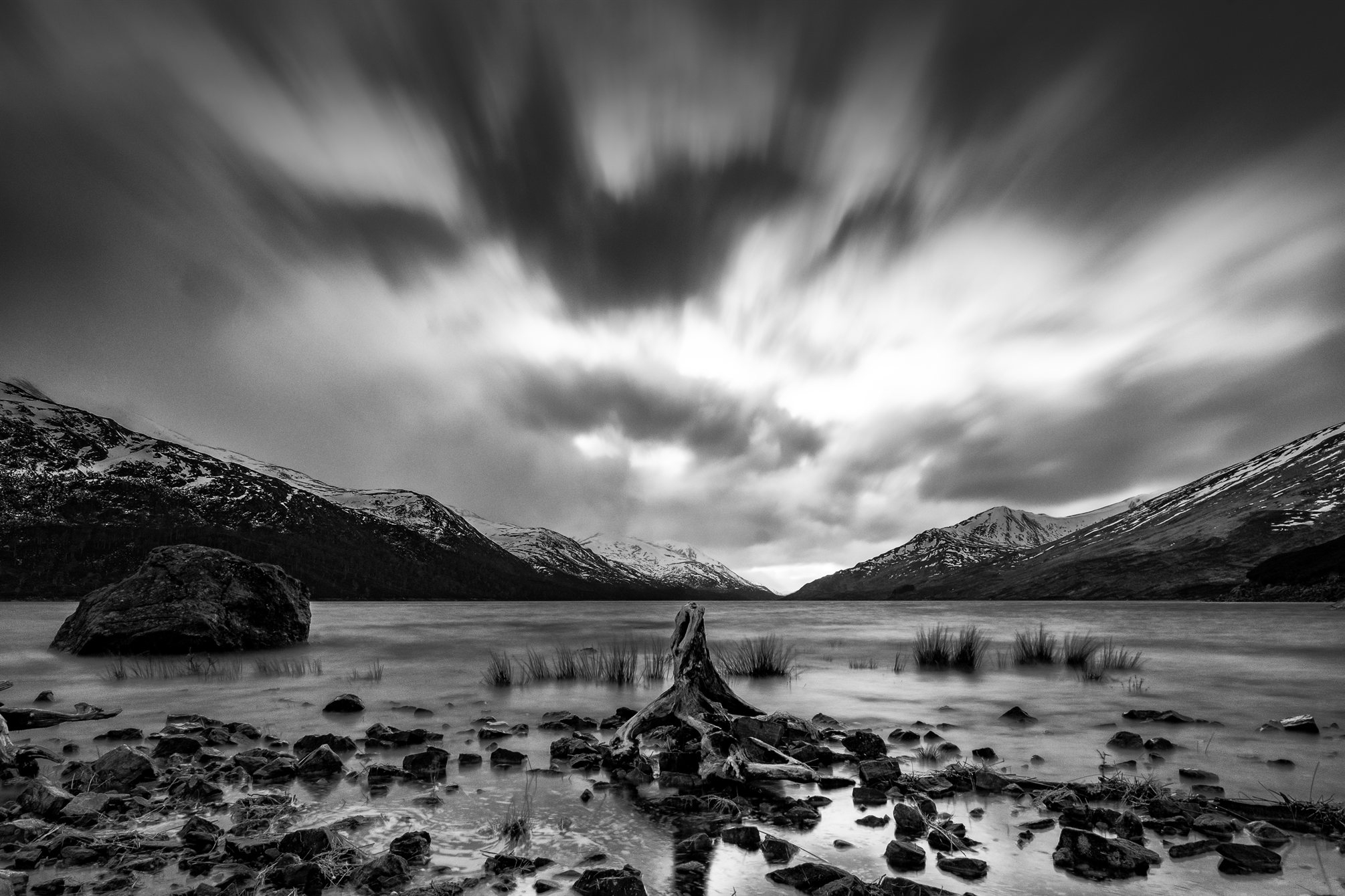 Snow-capped mountains around a loch under streaming clouds, with an old tree stump in the shallows, in black and white