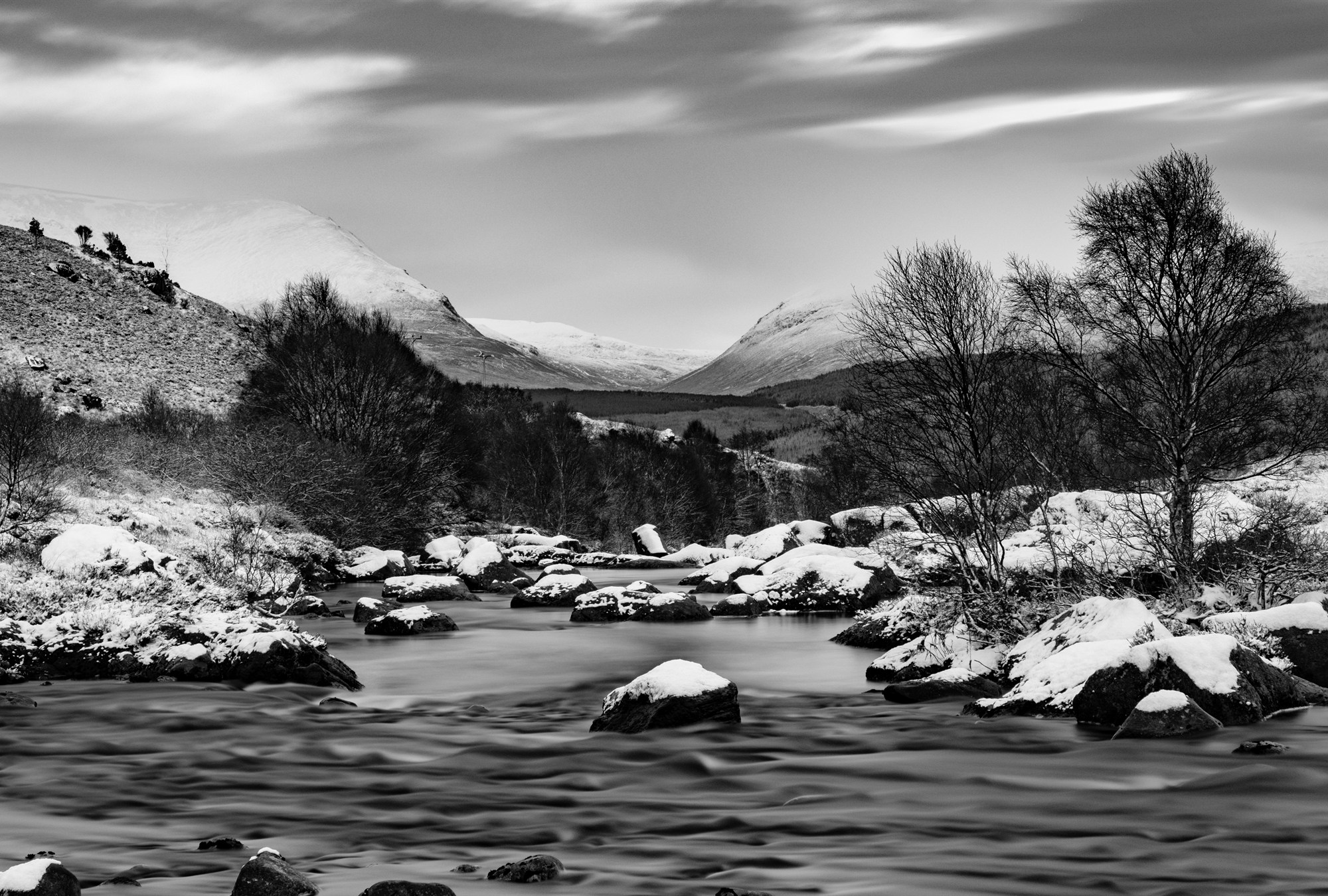 A rocky river dusted with snow, running towards snow-covered mountains, in black and white